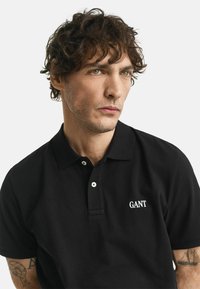 Man met krullend haar die een zwart GANT poloshirt draagt, kijkend naar de zijkant, met zichtbare tatoeages op zijn armen tegen een effen achtergrond.