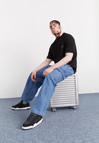 Ung man med vitiligo sitter på en silverfärgad resväska med hjul, iklädd en svart t-shirt, blå cargobyxor och svarta sneakers på en grå matta.