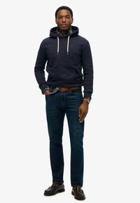 Sudadera azul marino con un logo texturizado, cordones beige, superpuesta sobre una camisa de cuadros. Combinada con jeans azul oscuro y mocasines marrones.