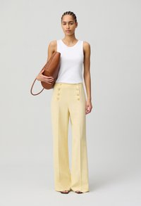 Pantalons taille haute en mélange de lin, de couleur jaune pastel, avec des accents de boutons à l'avant. Haut sans manches blanc. Modèle tenant un sac marron décontracté.