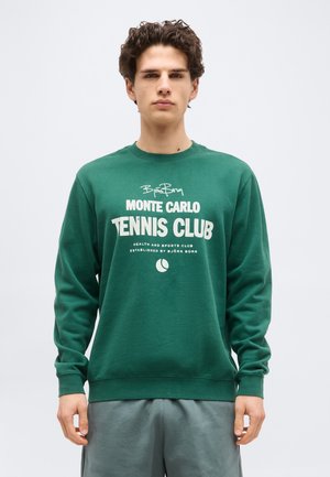 Νέος άνδρας με σκούρα σγουρά μαλλιά, φορώντας πράσινο φούτερ με το κείμενο "Monte Carlo Tennis Club" και γραφικό με μπάλα του τένις, στέκεται μπροστά σε λευκό φόντο.