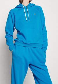 Person som bär en ljusblå hoodie och matchande sweatpants med händerna i fickorna mot en enfärgad bakgrund.