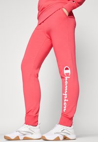 Pantaloni de trening corali cu un design fit, buzunare laterale și logo-ul alb "Champion" pe piciorul stâng. Asortați cu încălțăminte sport albă.