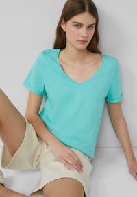 Turkusowa koszulka V-neck wykonana z miękkiej bawełny, z krótkimi rękawami i podwiniętymi mankietami, w komplecie z beżowymi szortami, na neutralnym tle.
