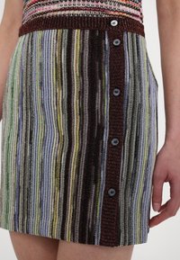 Sukienka dzianinowa z wielokolorowymi pionowymi paskami o różnych fakturach, z bordowym pasem w talii i sześcioma czarnymi guzikami po boku.