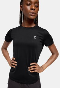 T-shirt de sport noir avec un col rond, des manches courtes et un petit logo blanc sur la poitrine gauche. Tissu léger et texturé.