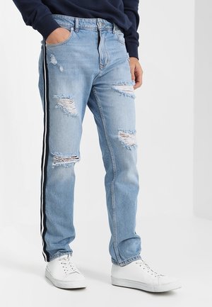 Lichtblauwe gescheurde spijkerbroek met zwart-witte zijstrepen, gedragen met witte sneakers en een marineblauw shirt met lange mouwen.