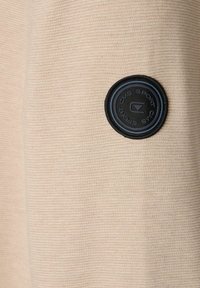 Beżowy materiał na bluzę z textured żeberkowym wzorem. Posiada czarną okrągłą metkę z wytłoczonym napisem "CMS Sport" pośrodku.
