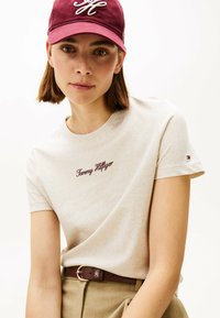 Beige bomulds t-shirt med runder hals, der har maroon "Tommy Hilfiger" logo broderi. Parret med et brunt bælte og en bourgogne kasket.