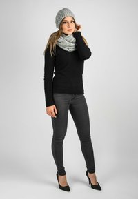 Un bonnet en tricot gris et une écharpe assortie, un pull noir ajusté, un jean foncé et des talons hauts noirs avec des clous en argent, se tiennent devant un fond neutre.