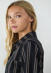 Schwarz gestreiftes Hemd mit braunen und weißen Linien, mit Kragen und einer vorderen Tasche, aus einem glatten Stoff gefertigt.