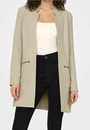 Vrouw die een beige blazer met open voorkant en ritszakken draagt, een wit topje met vierkante hals en zwarte broek met hoge taille.