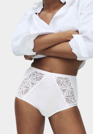 Vrouw draagt witte hoge tailleonderbroek met kanten inzetstukken en een wit overhemd met opgerolde mouwen, armen gekruist over de borst.