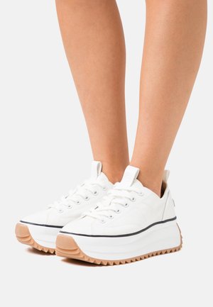 Zapatillas de lona blancas con suela de goma gruesa y texturizada, línea de acento negro y cierre de cordones en la parte delantera. Tirador en el talón para facilitar su colocación.