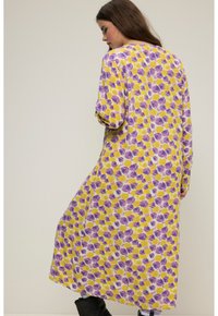 Vestido largo y holgado hecho de una tela ligera, con un patrón floral amarillo y morado. Escote en V y mangas largas con un dobladillo redondeado.