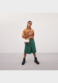 Brązowy t-shirt z nadrukiem, zielone szorty, czarne buty i kremowa torebka na łańcuszkowym pasku. Tło studyjne w neutralnych odcieniach.