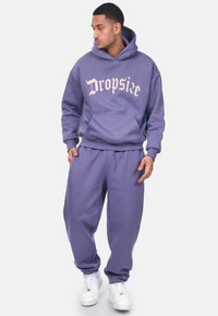 Sudadera morada y pantalones de chándal a juego fabricados en una tela suave. Presenta un bolsillo frontal y un gran detalle de texto en rosa claro. Se llevan zapatillas blancas.
