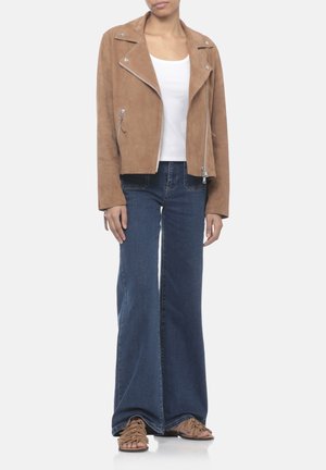 Donna che indossa una giacca in camoscio marrone, top bianco, jeans larghi blu e sandali marroni intrecciati, in piedi davanti a uno sfondo bianco semplice.