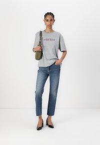 Šedé tričko s červeným logem "GUESS JEANS", spárované s modrými rovnými džíny s třepením na lemu a černými lakovanými vysokými podpatky.