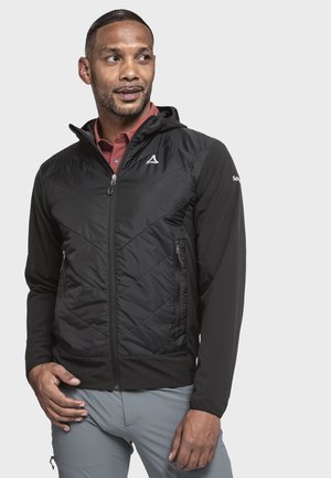 Mann trägt eine schwarze gesteppte Kapuzenjacke über einem roten Hemd und hellgrauen Hosen, blickt leicht zur Seite mit neutralem Gesichtsausdruck.