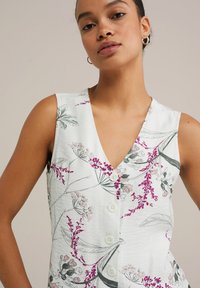 Mouwloze bloemenjurk van witte stof met roze en groene botanische patronen, met een diepe V-hals en knopen aan de voorkant.