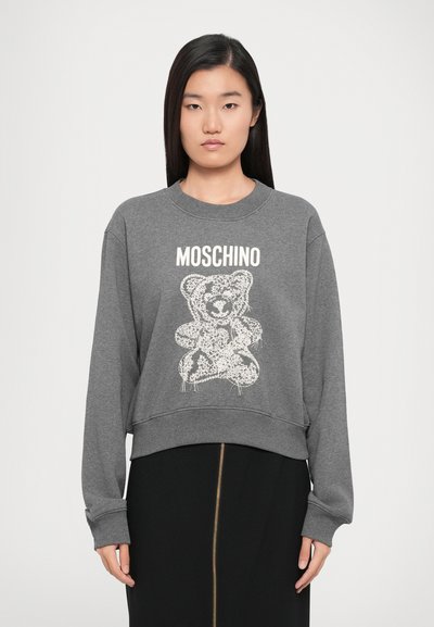 Szara bluzka z kapturem o skróconym kroju, z długimi rękawami, ściągaczami przy mankietach oraz grafiką białego niedźwiedzia. Napis "Moschino" znajduje się nad wzorem.