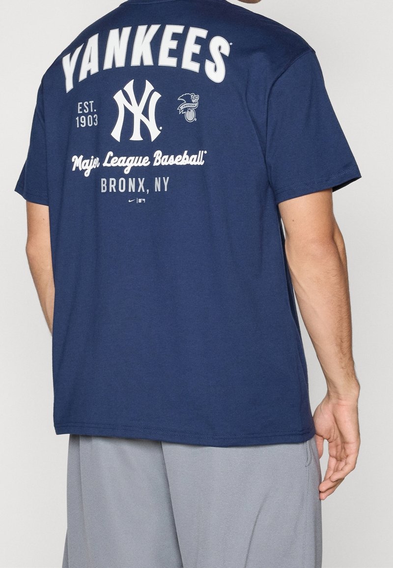 Homme portant un T-shirt bleu marine des Yankees avec texte et logos blancs, associé à un short gris clair, vu de dos.