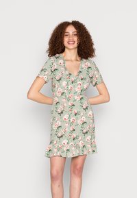 Vestido curto floral em verde claro, com decote em V, mangas curtas e bainha com babados, adornado com padrões de flores em rosa e branco.