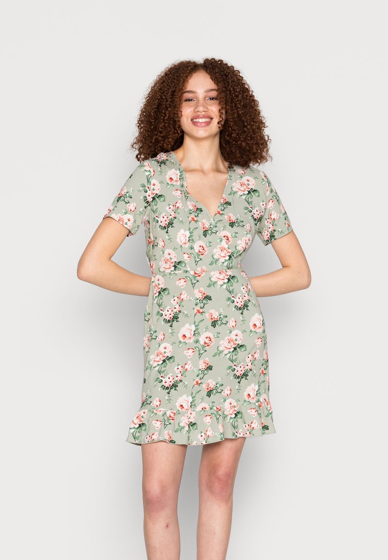 Vestido curto floral em verde claro, com decote em V, mangas curtas e bainha com babados, adornado com padrões de flores em rosa e branco.