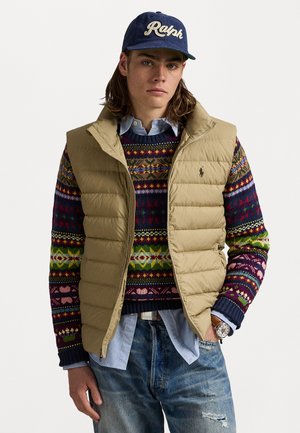 Young man wearing a beige quilted vest over a colorful patterned sweater, denim shirt, distressed jeans, blue cap, and a watch with a brown leather strap.