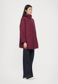 Veste à capuche bordeaux avec fermeture éclair, design structuré et coupe ample. Associée à un jean en denim foncé et des chaussures blanches à bout pointu.