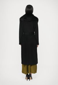 Melnas vilnas mētelis ar mākslīgās kažokādas apkakli, jostu viduklī un garu siluetu. Valkāts kopā ar zaļu svārku, papildināts ar melnām augstpapēžu kurpēm.