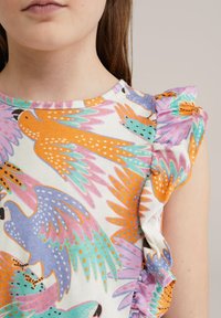 Camiseta de algodón colorida con un loro y un patrón floral, con cuello redondo y acentos de hombros con volantes.