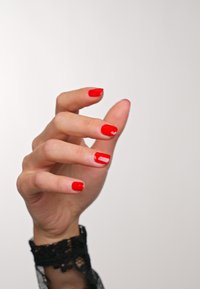 Mano con unghie laccate di rosso, dita leggermente ricurve. Polsino in pizzo visibile al polso. La pelle liscia e il colore vivace contrastano con uno sfondo chiaro.