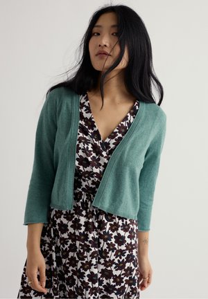 Joven con cabello largo y negro, vistiendo un cárdigan azul verdoso sobre un vestido blanco con patrones florales oscuros, de pie frente a un fondo liso.