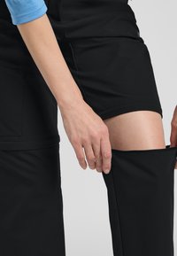 Zwarte zachte stoffen shorts met een ritsgedeelte. Een hand houdt de zoom vast, waardoor een gladde, ononderbroken kledinglijn zichtbaar is. Blauwe mouw zichtbaar.