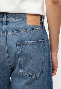 Primo piano della vita di una persona con jeans di denim blu, toppa del marchio in pelle marrone e camicia bianca infilata.