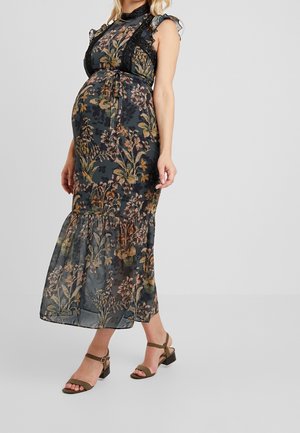 Robe longue en tissu sombre à motifs floraux, avec des épaules à volants, des détails en dentelle et un ourlet à plusieurs niveaux. Associée à des talons blocs vert olive.