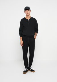 Crna dugoruka henley majica s detaljima gumba, u kombinaciji s crnim tapered jogger hlačama s džepovima, i crno-bijelim niskim sportskim tenisicama.