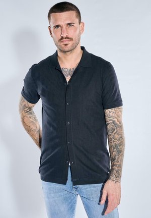 Mann mit kurzen Haaren und Tätowierungen an beiden Armen steht in einem schwarzen, kurzärmeligen Hemd mit Knöpfen und hellblauen Jeans vor einem einfarbigen Hintergrund.