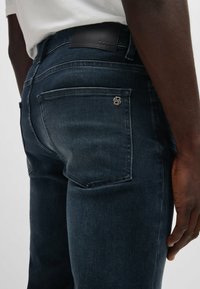 Dark blue denim jeans with a smooth texture. Features a leather waistband label and a small metallic logo on the back pocket.