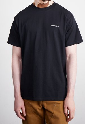 Person iført en sort Carhartt T-shirt med korte ærmer og brune bukser, stående mod en ensartet lys baggrund.