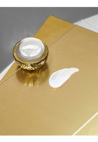 Crema antienvejecimiento blanca en un tarro redondo de oro texturizado, con una textura cremosa y suave, sobre una superficie dorada reflectante, con crema derramada a su lado.