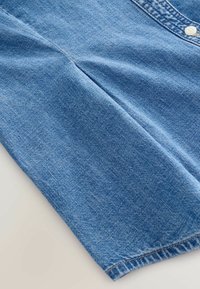 Denimskjorte i blå farve med knaplukning, krave og en glat overflade, der har synlige stikninger langs nederste kant og placket.