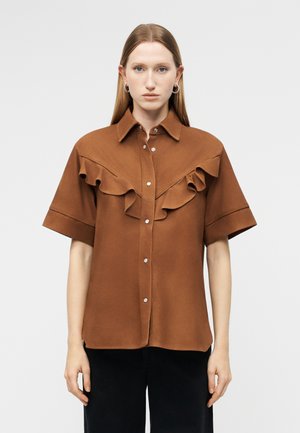 Γυναίκα με ίσια μαλλιά, που φοράει καφέ κοντομάνικο πουκάμισο με βολάν στο στήθος και ασημένια κουμπιά, σε συνδυασμό με μαύρο παντελόνι, μπροστά σε απλό φόντο.