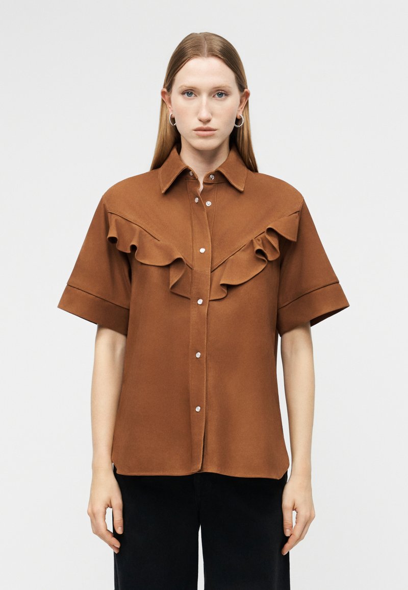 Mujer con cabello lacio que lleva una camisa marrón de manga corta con canesú con volantes y botones plateados, combinada con pantalones negros, sobre un fondo liso.