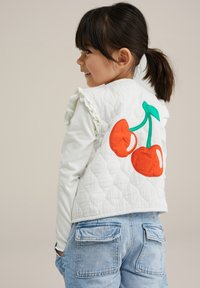 Vadderad vit väst med volangkrage, med en design av orange körsbär på baksidan. Kombinerad med ljusblå denimjeans.