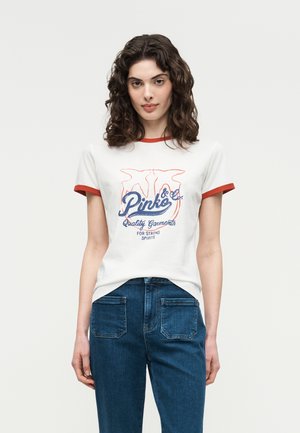 Žena s kučeravými vlasmi, oblečená v bielom tričku s červeným lemom a modrým nápisom, v modrých džínsoch, stojaca pred jednofarebným svetlým pozadím.