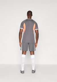 Grå idrettsdrakt med oransje detaljer, kombinert med matchende shorts. Modellen har på seg hvite sokker og grå fotballsko, som viser et sporty design.