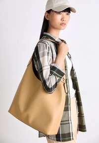 Jeune femme portant une casquette beige et une chemise à carreaux, tenant un grand sac fourre-tout beige sur l'épaule, sur un fond uni.